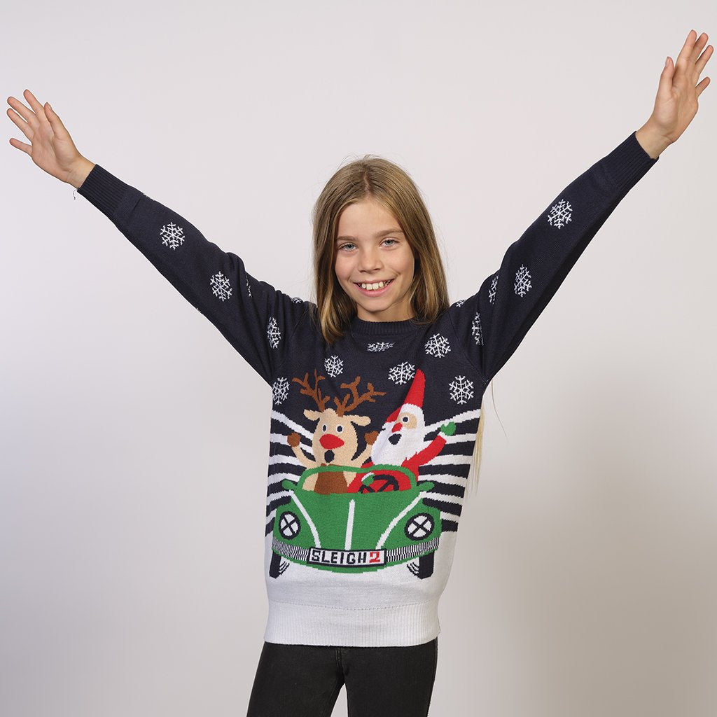 Un enfant posant les bras en l'air portant un pull bleu de Noël avec Rudolph et le Père Noël dans une voiture.