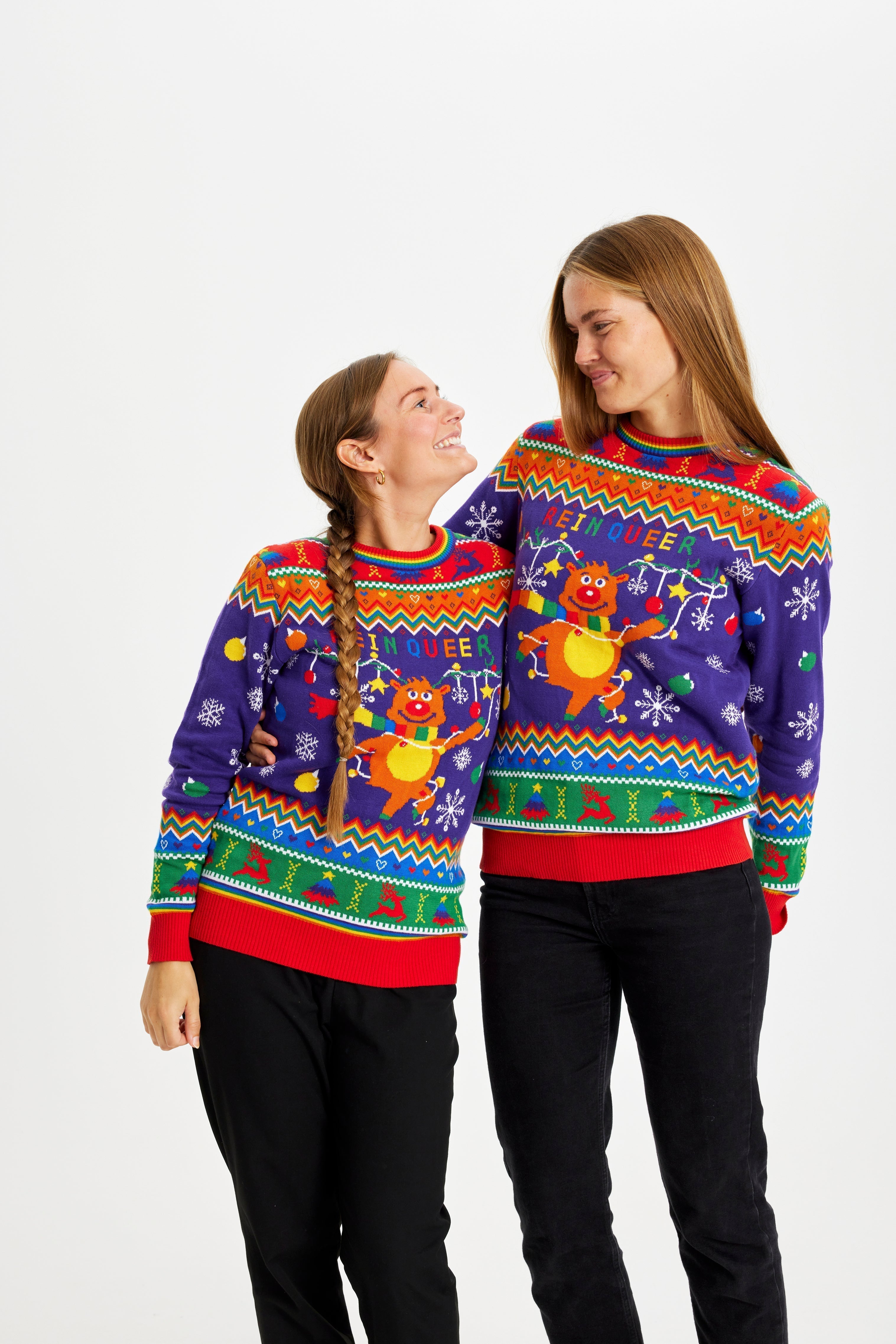 Deux femmes se regardant l'une l'autre et portant un pull de Noël aux couleurs de l'arc-en-ciel avec la citation 