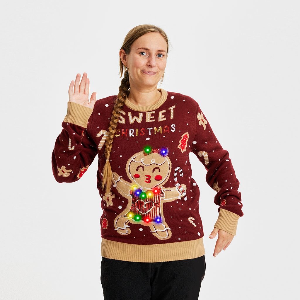 En smilende dame, der vinker, står i en mørkerød julesweater med en kageperson og lys på.