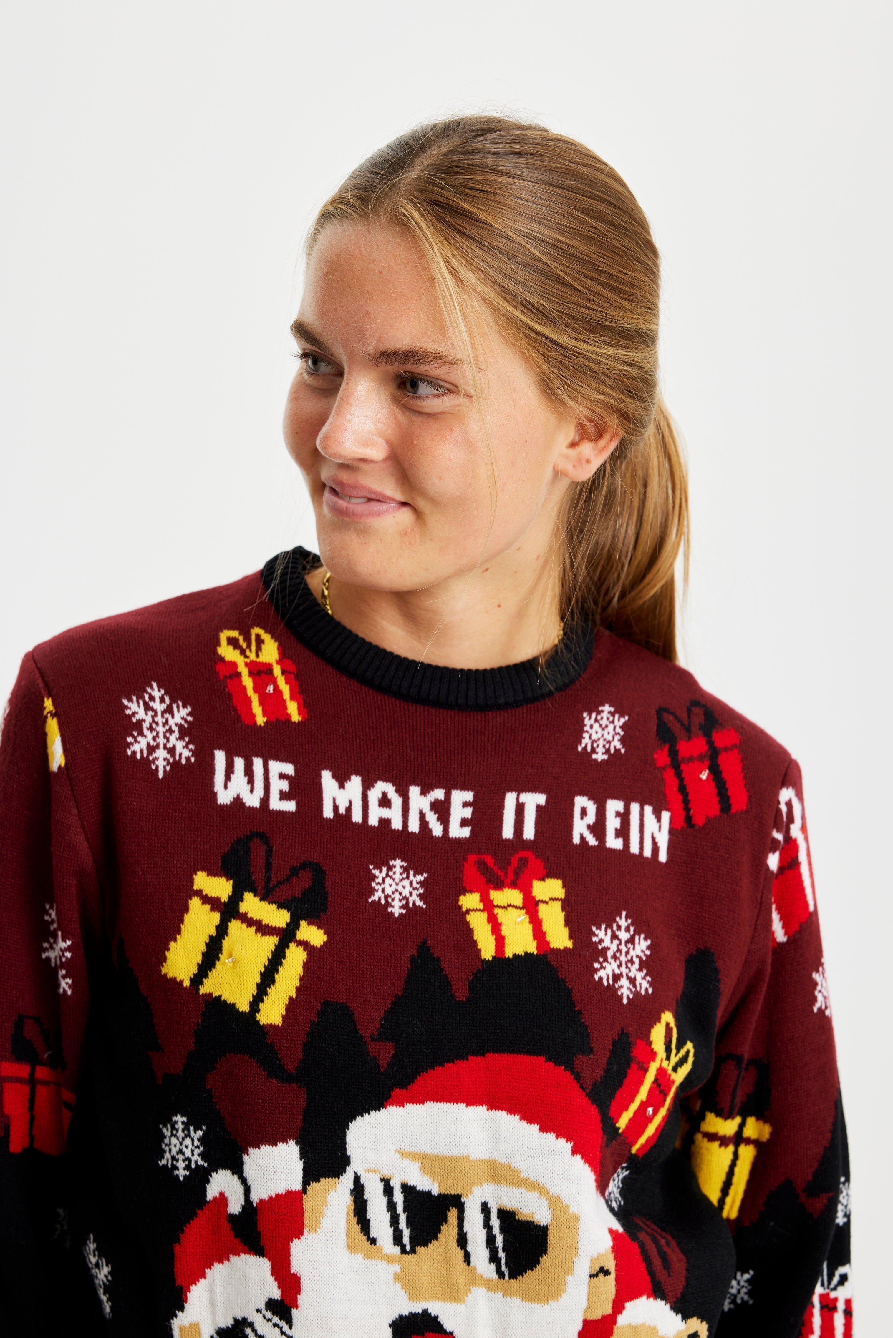 Une femme qui pose regarde sur le côté, vêtue d'un pull de Noël rouge et noir avec le Père Noël et des lumières.