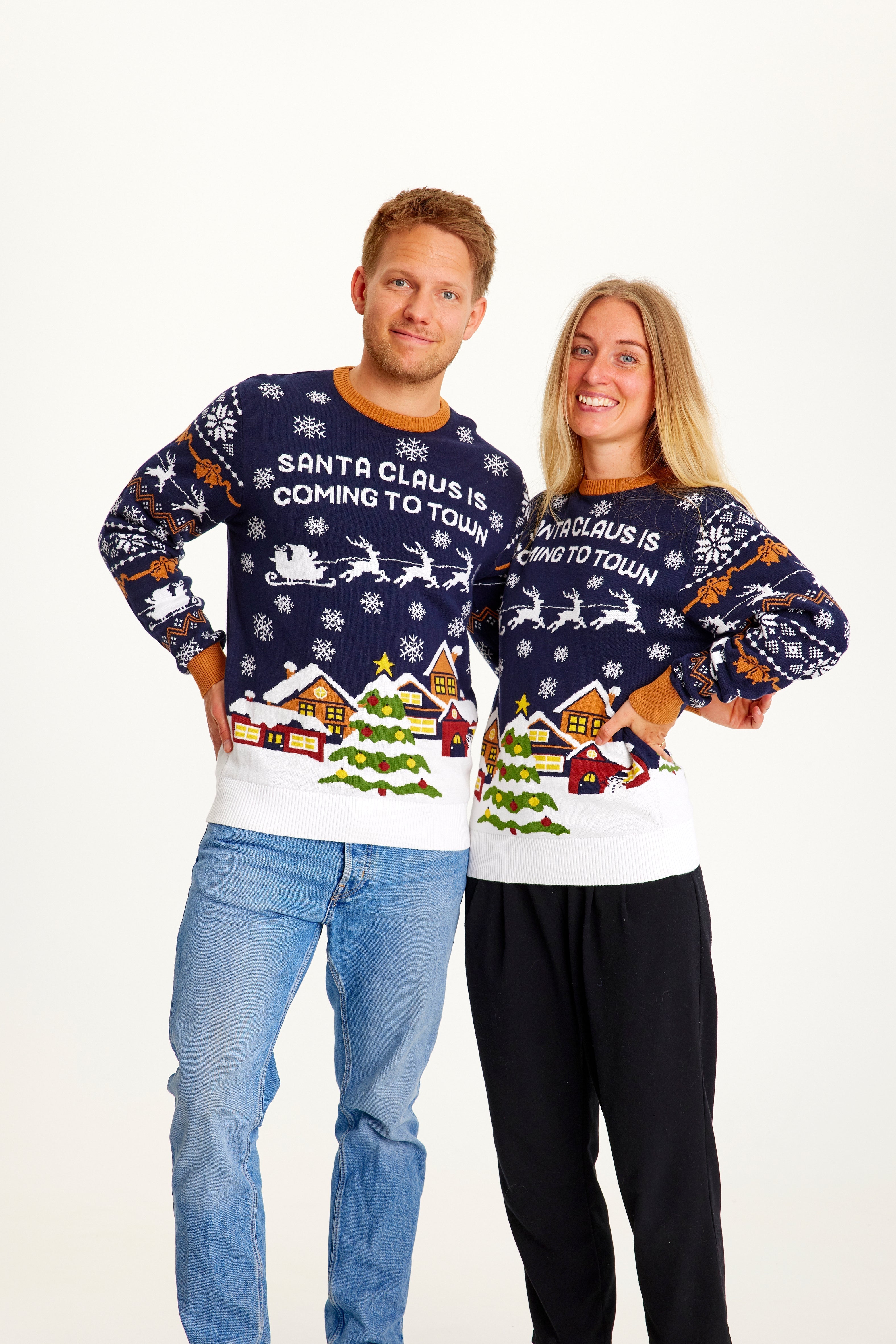 Un couple, les mains le long du corps, portant un pull de Noël bleu foncé et blanc orné d'un arbre de Noël.